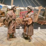 Figuras de personas en una escena de comercio antiguo junto a un barco.