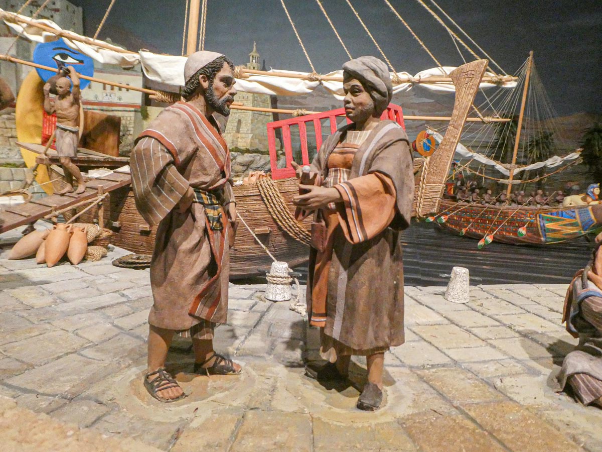 Figuras de personas en una escena de comercio antiguo junto a un barco.