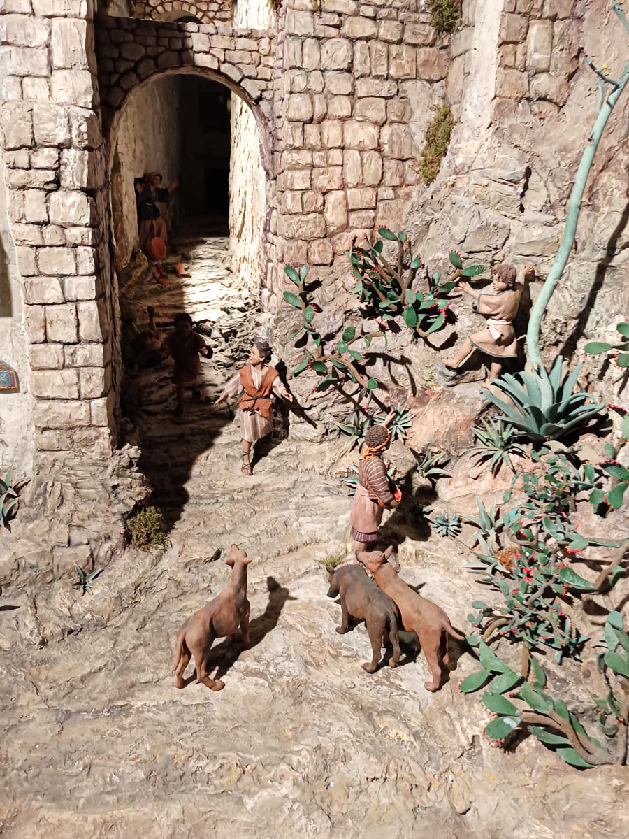 Escena de una calle con figuras humanas y perros en un paisaje rocoso.