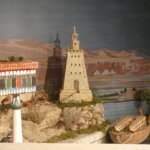 Modelo de un puerto antiguo con un faro y barcos en un paisaje montañoso.