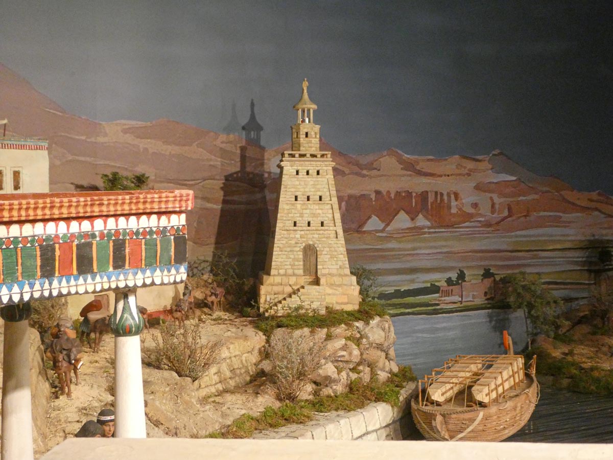 Modelo de un puerto antiguo con un faro y barcos en un paisaje montañoso.