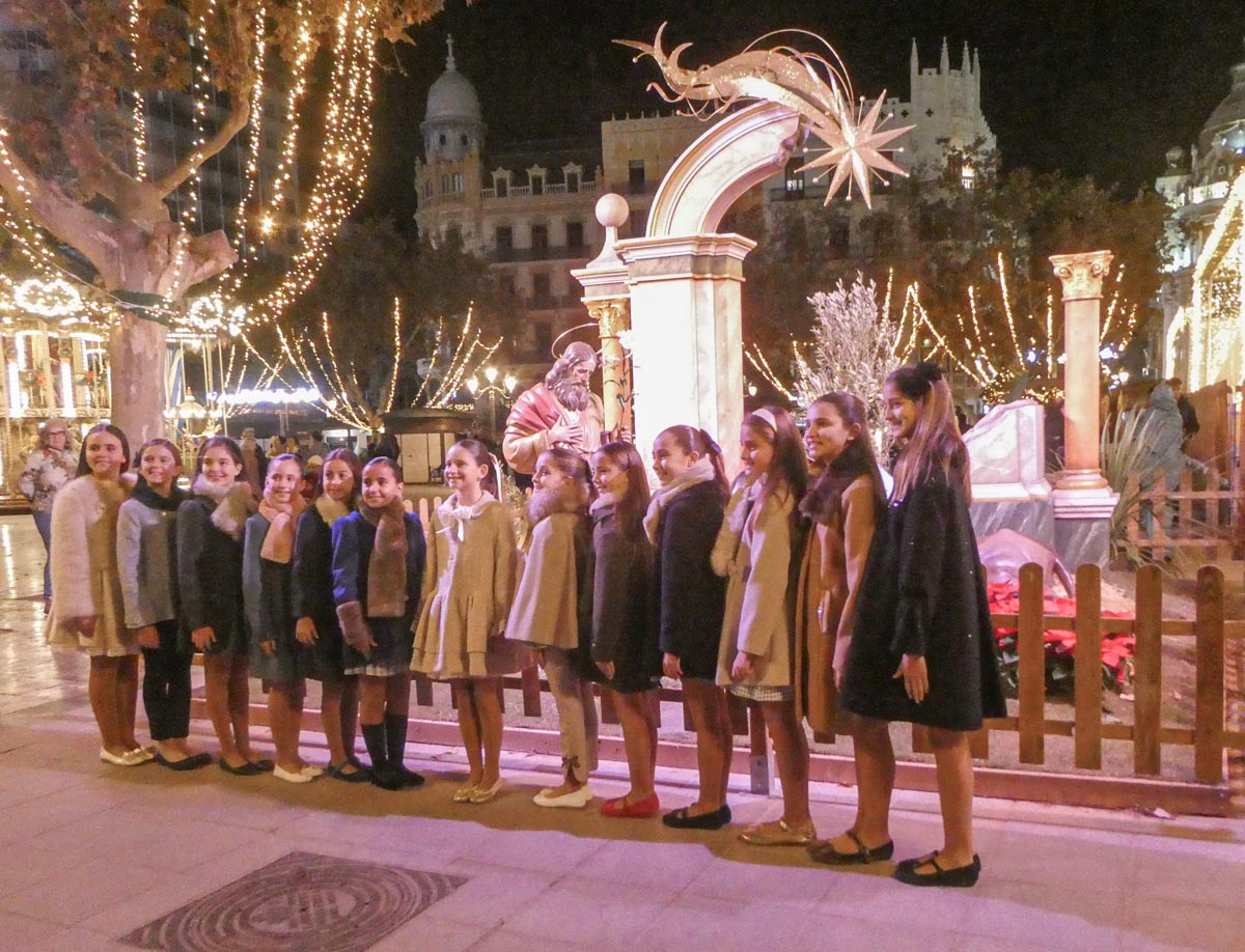 Grupo de niñas posando en una plaza iluminada por luces navideñas.