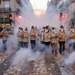 Grupo de personas con mascarillas en un desfile con humo y fuegos artificiales.