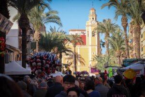 Multitud de personas en una calle con palmeras y un campanario amarillo