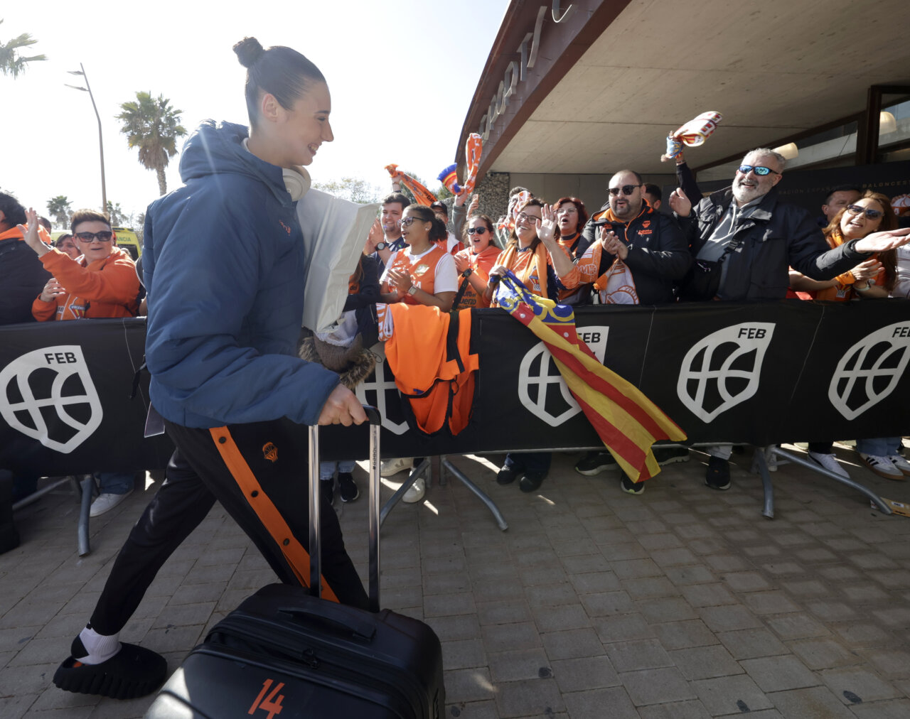 Jugadora de Valencia Basket llegando con maleta mientras aficionados celebran.