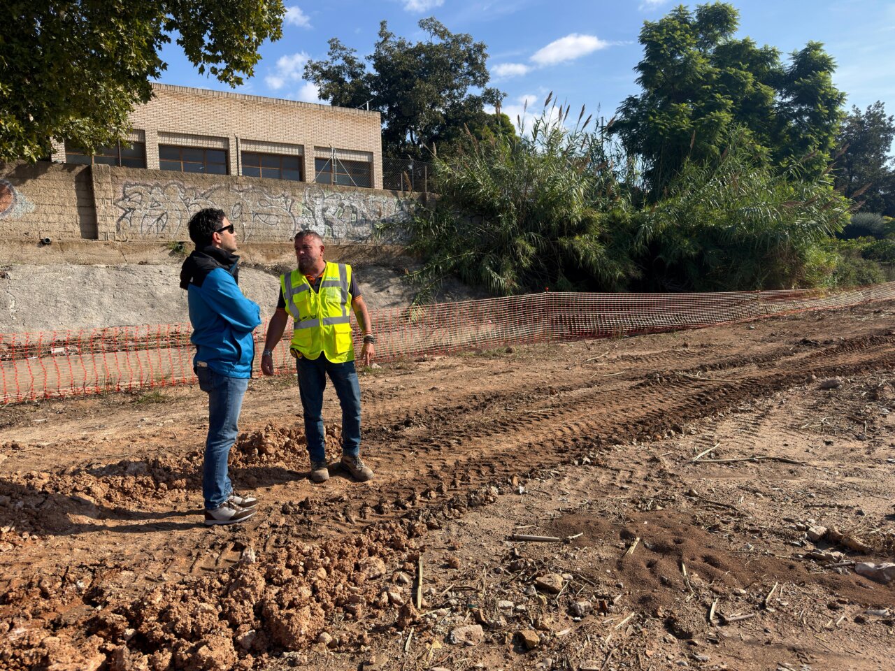 Dos hombres junto a terreno removido, con una malla naranja delimitando la zona de obras.